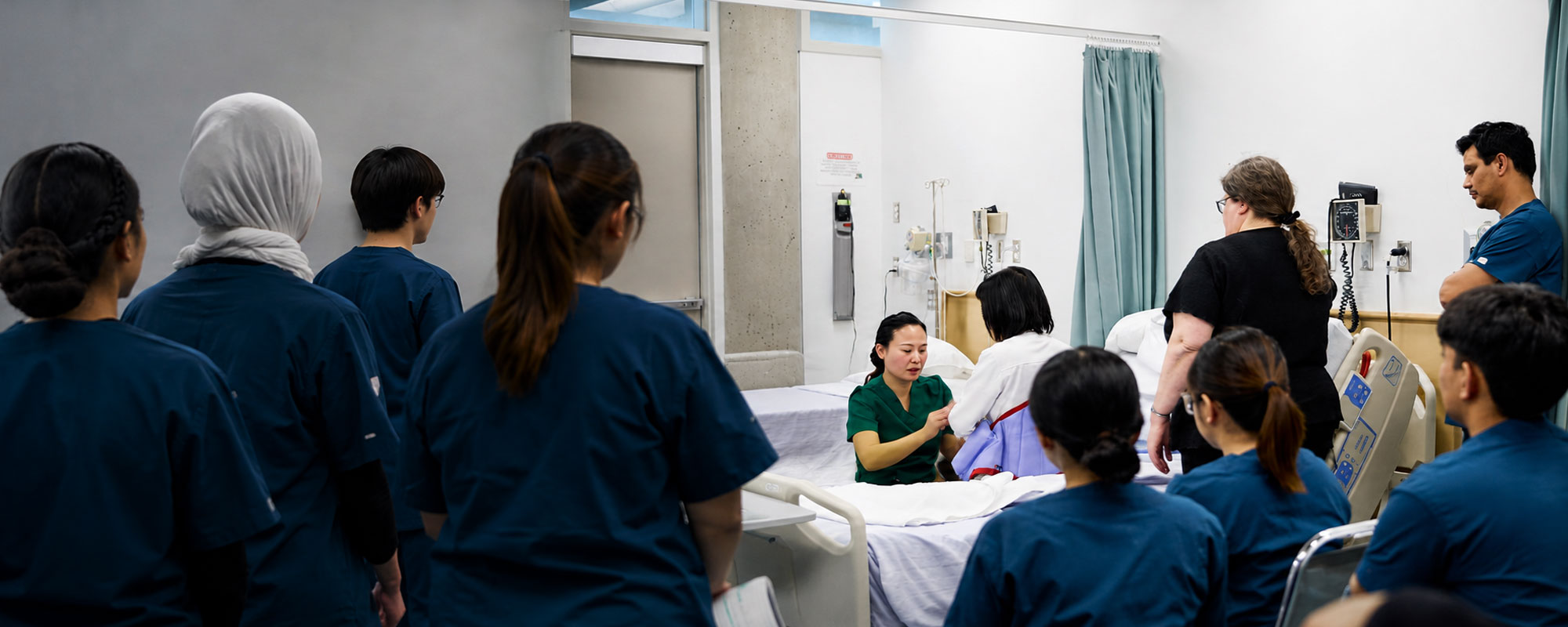 A group of healthcare assistant students in scrubs gather in a clinical training room to watch a demonstration of a mechanical patient lift being operated by an instructor.