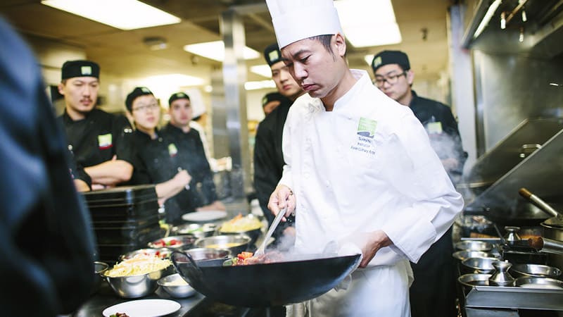 Chef in a white uniform and hat focuses intently on cooking with a wok in a busy kitchen. Students in dark uniforms watch attentively.