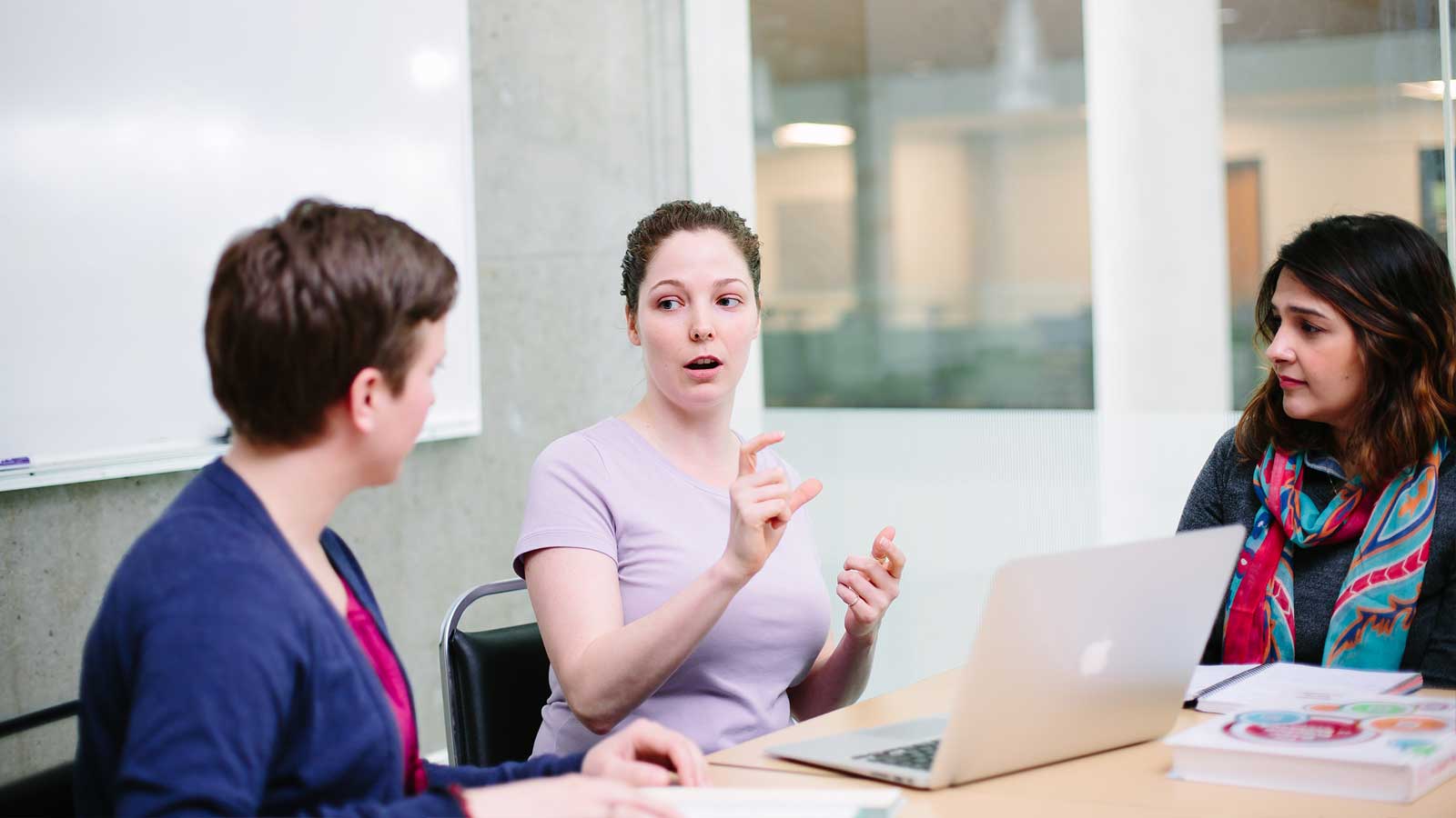 Three people sit around a table in discussion. A woman in the center gestures while speaking, with a laptop and books visible.