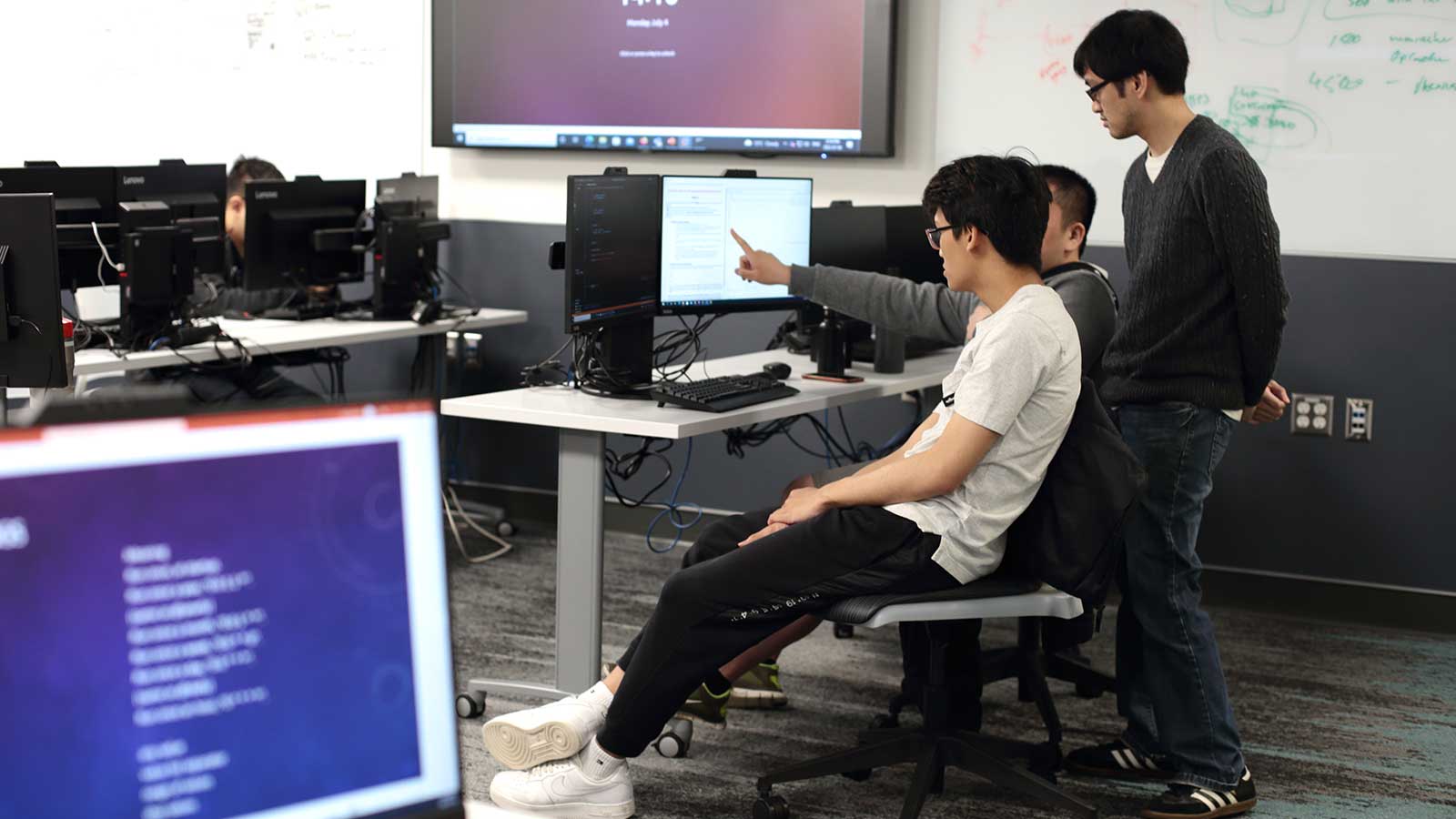 Three men collaborate on computers in a classroom, surrounded by educational materials and a learning environment.