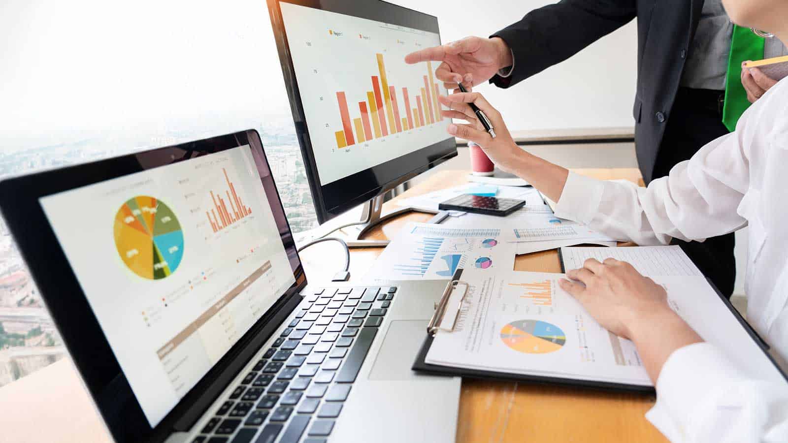 Two people analyzing graphs on a laptop and monitor, pointing at charts with documents laid out on the desk.