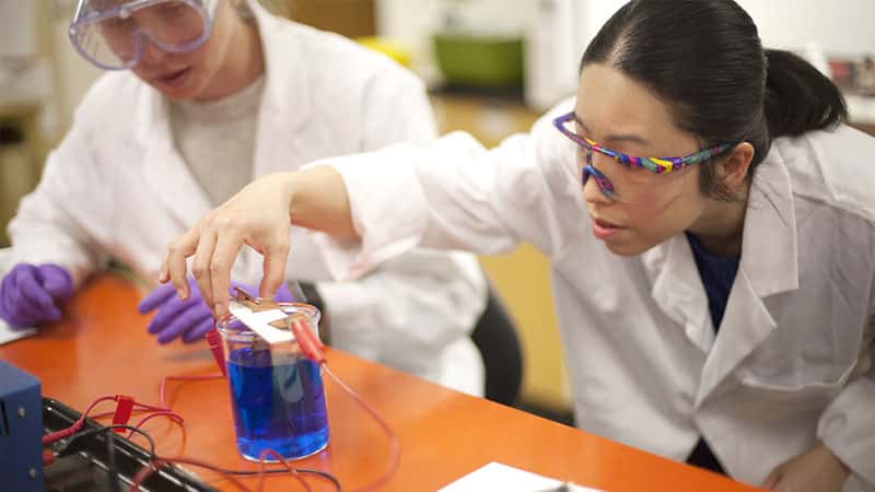 Two scientists in lab coats and safety goggles conduct an experiment. One adjusts a device in a beaker of blue liquid.