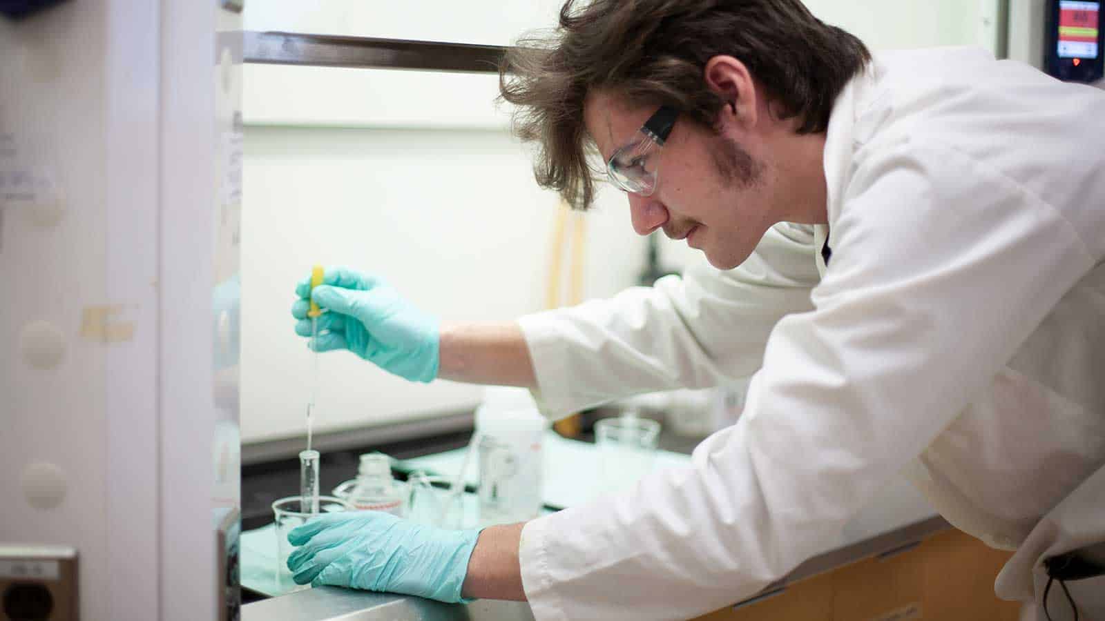 A scientist wearing a lab coat, safety glasses, and gloves uses a pipette in a laboratory.