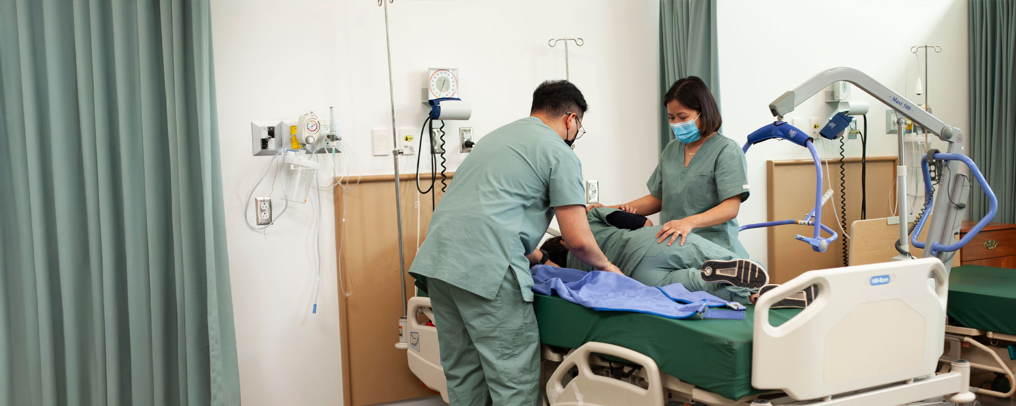 Two nursing students wearing green scrubs and face masks safely reposition a patient on a hospital bed in a training ward.