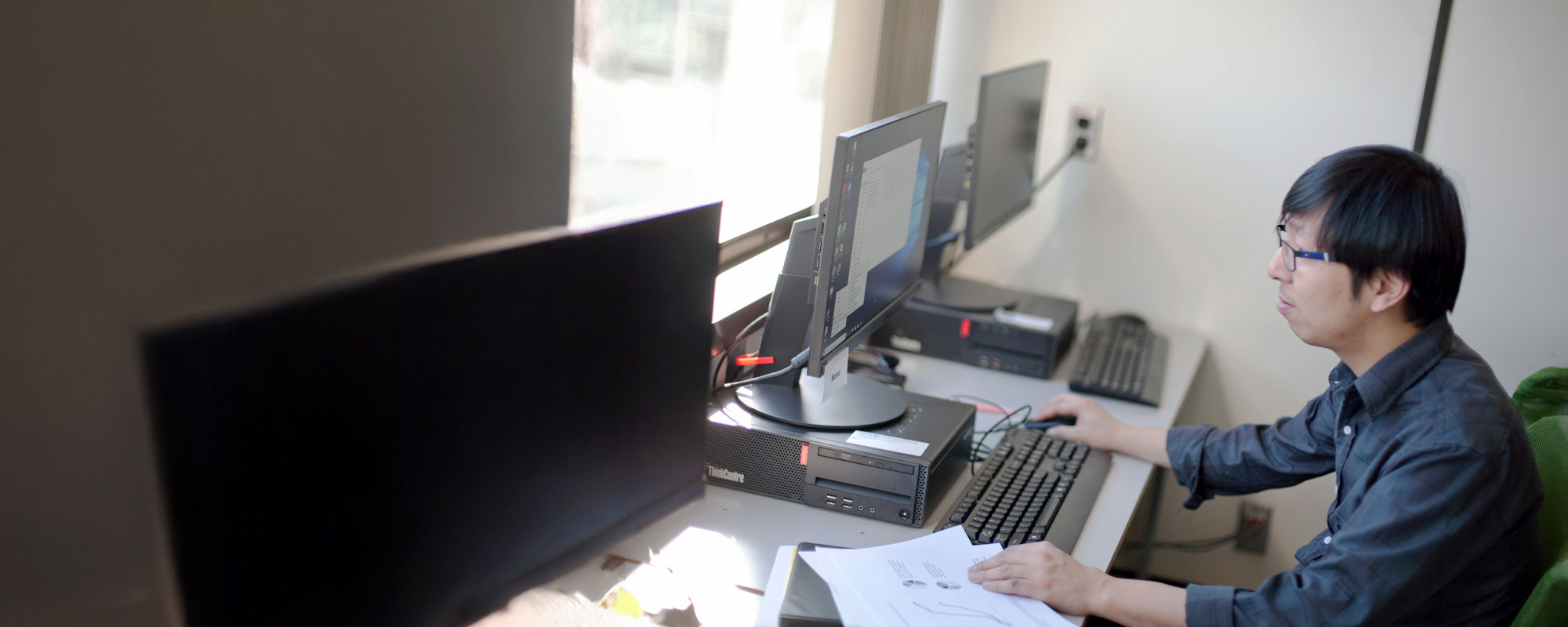 a student wearing glasses works at a computer terminal while reviewing printed financial documents and charts.
