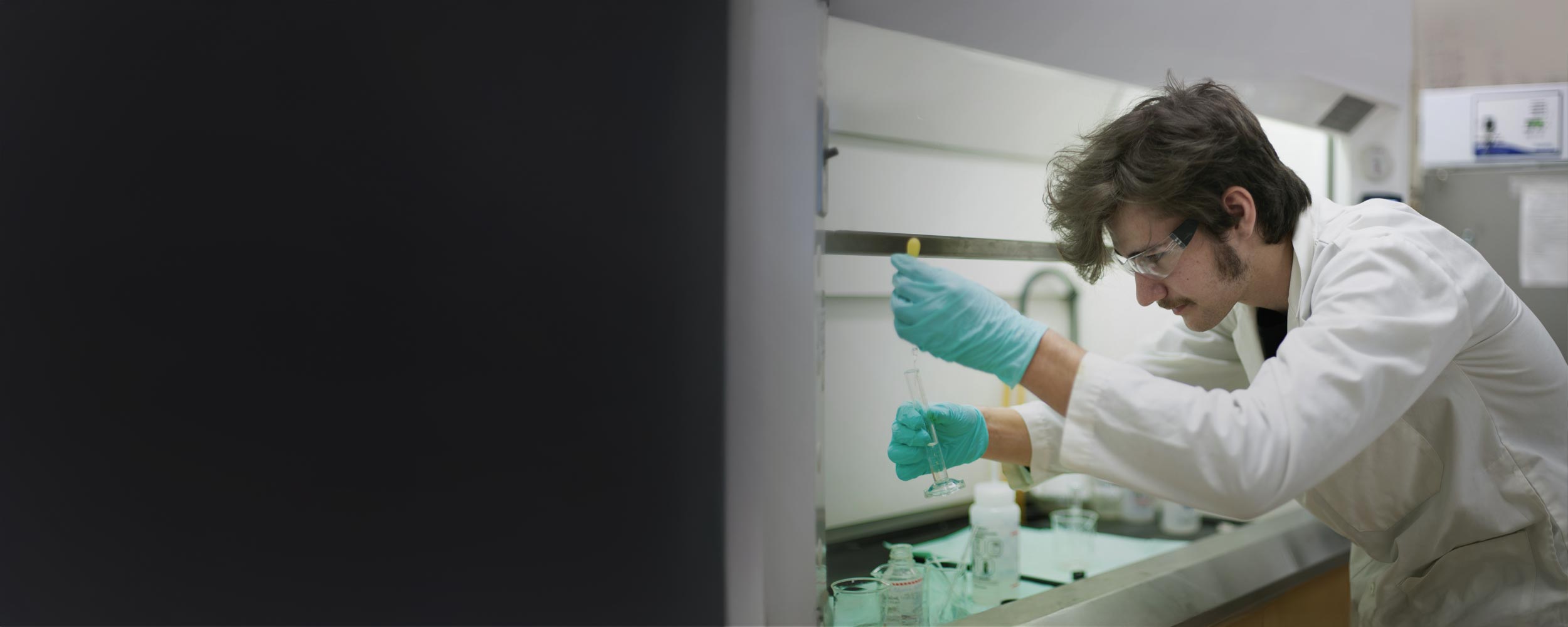 A student researcher wearing safety glasses, a white lab coat, and green protective gloves carefully transfers a liquid into a graduated cylinder inside a laboratory fume hood.