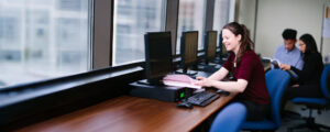 A smiling student reviews paperwork at a computer workstation in a bright office or lab while other students work in the background.