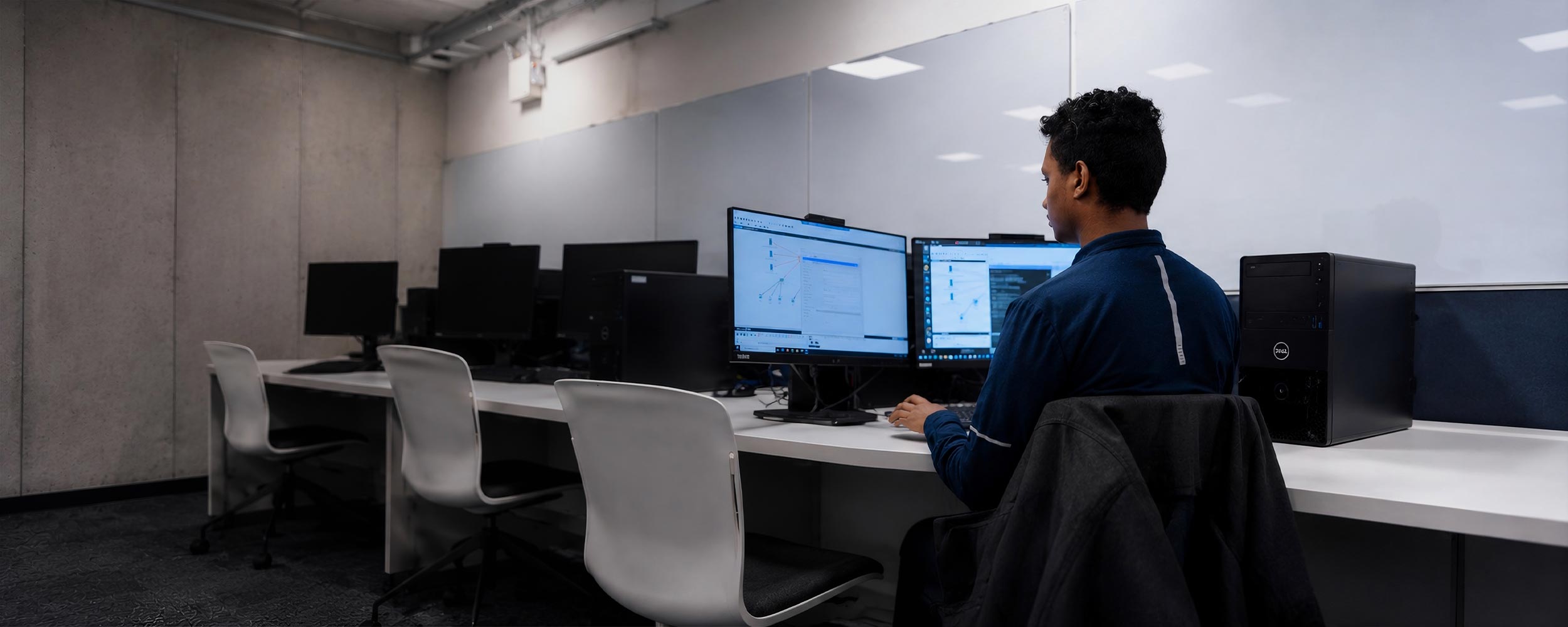 A student works at a dual-monitor workstation displaying network diagrams in a modern computer lab.