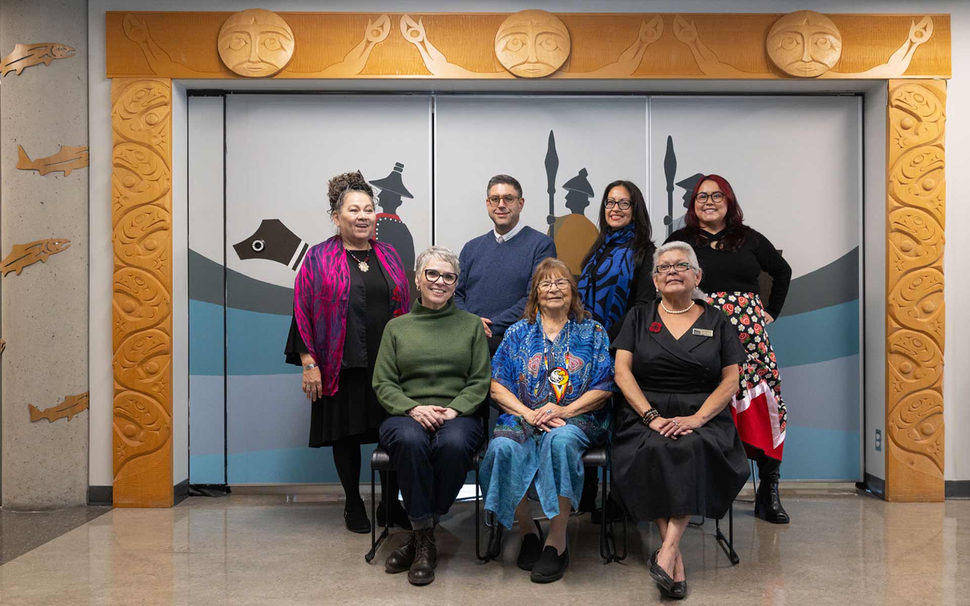 Indigenous elsers and counsellors gathered in a group portrait in front of a indigenous mural of a canoe