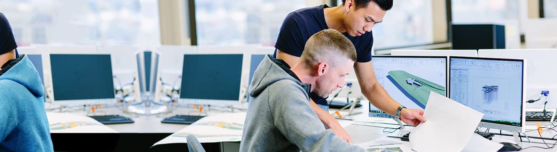 Two people collaborating at a desk with dual monitors displaying technical diagrams in a bright office setting.