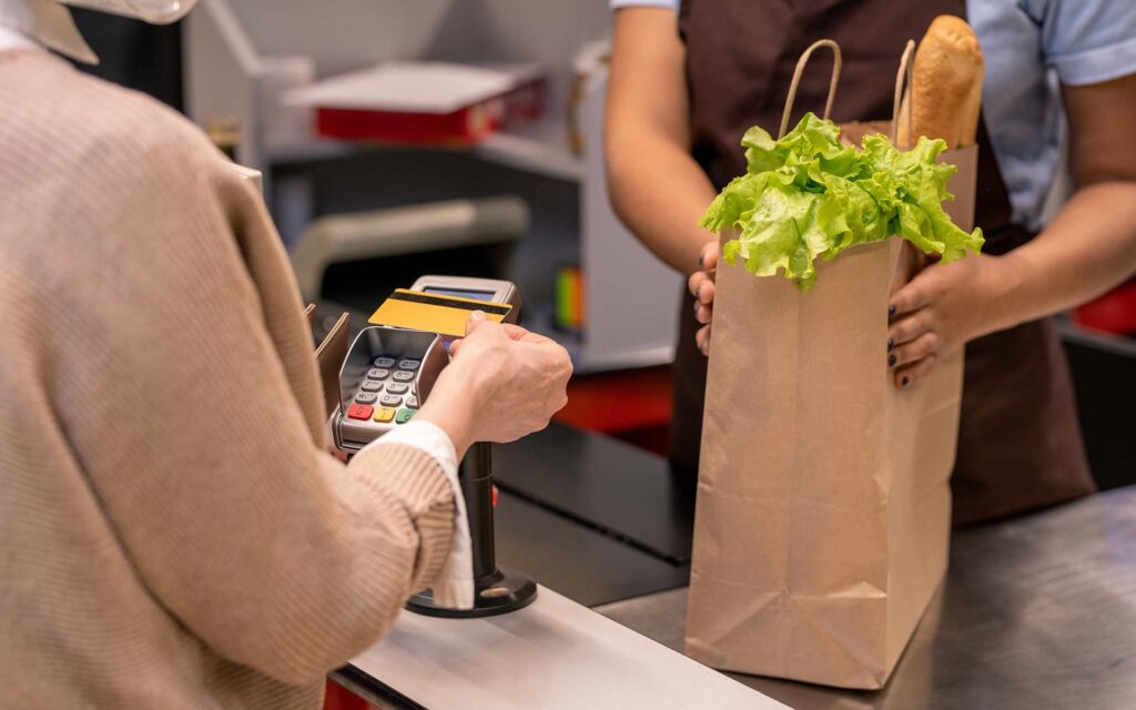 A person paying for groceries using a card reader at a checkout counter, with a cashier holding a bag of fresh lettuce and bread.