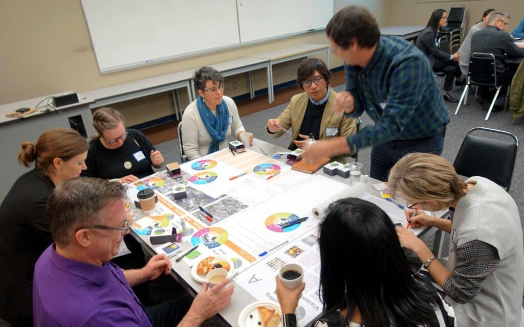 A diverse group of eight people are seated around a square table covered with papers, diagrams, and coffee cups, actively engaged in a lively group discussion indoors.