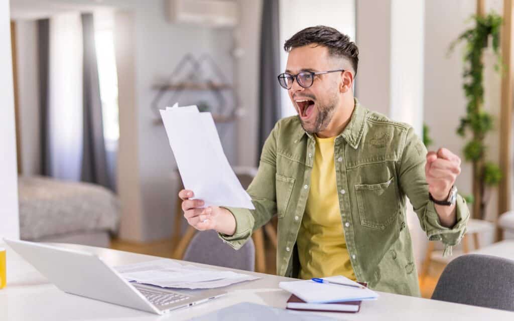 A man in a green jacket and yellow shirt, wearing glasses, sits at a table with a laptop and papers, excitedly cheering and raising his fist while holding up a document, with a home interior