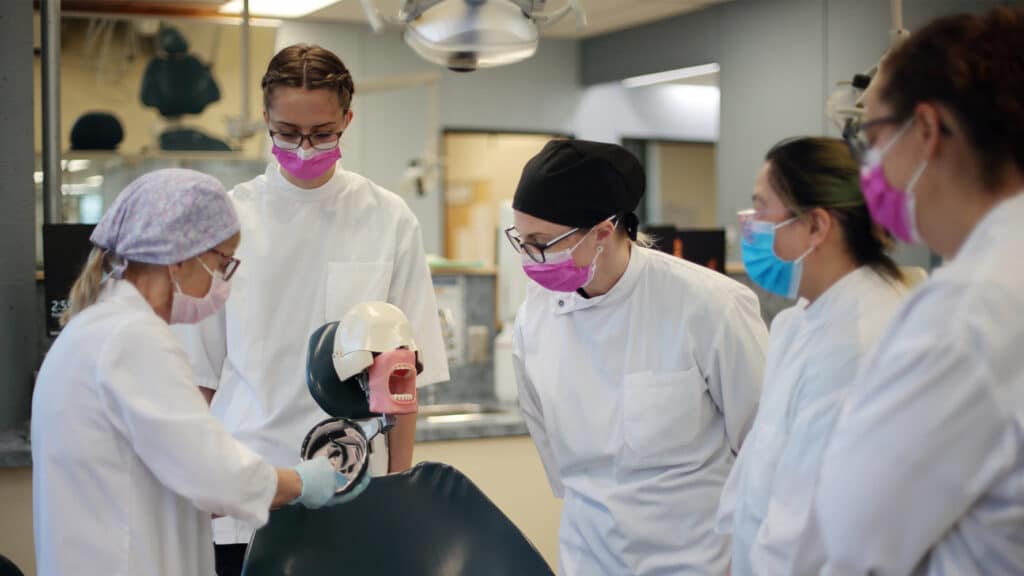 A dental instructor wearing blue gloves practicing a procedure on a dental training mannequin's mouth and teeth while dental students watch