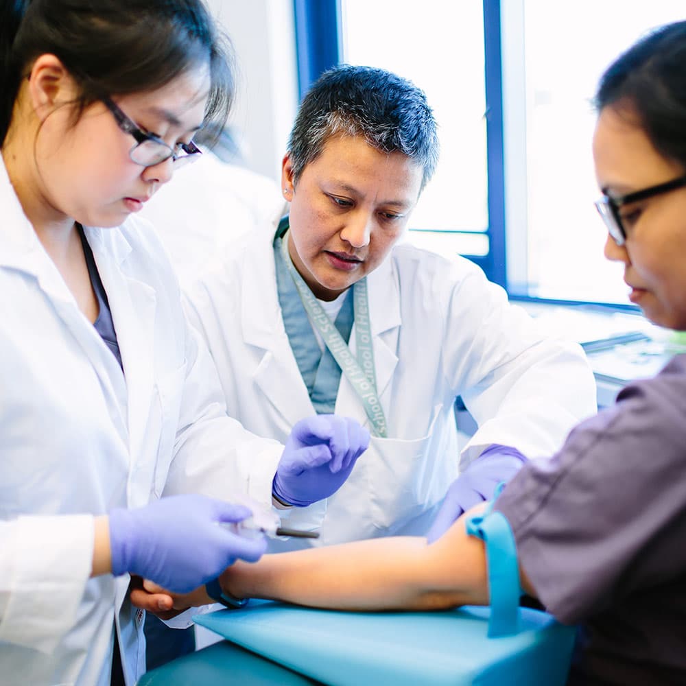 Medical lab assistants practicing a blood draw procedure on a person's arm, supervised by an instructor.