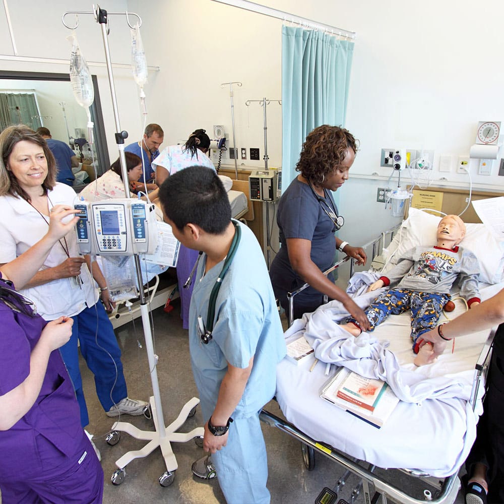 A group of nursing students and instructors practicing procedures like IV setup and pediatric care on dummies in a simulation ward.