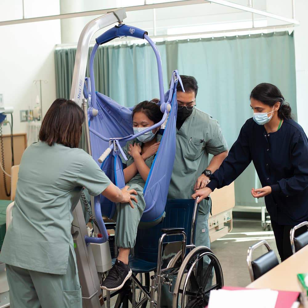 Nursing students using a mechanical patient lift to safely transfer a client from a wheelchair in a simulated clinical setting.