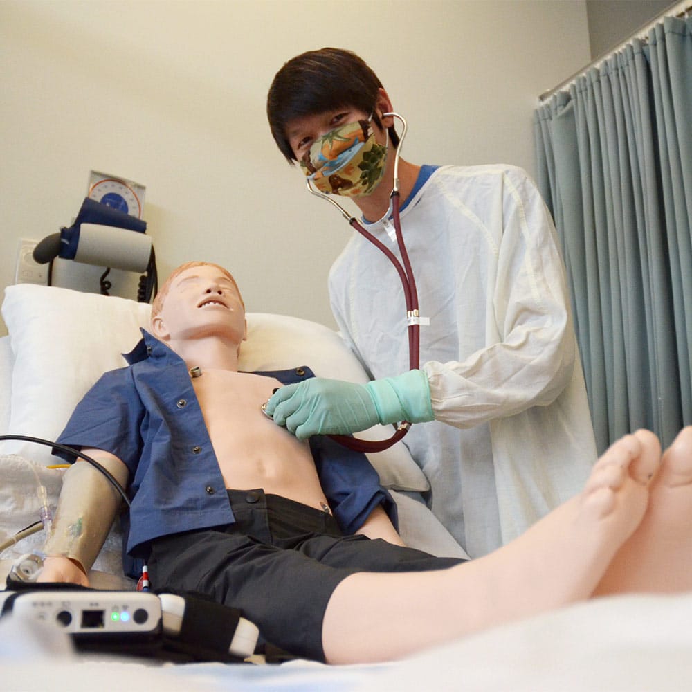 A healthcare student wearing a mask and gown using a stethoscope on a pediatric patient simulation child.