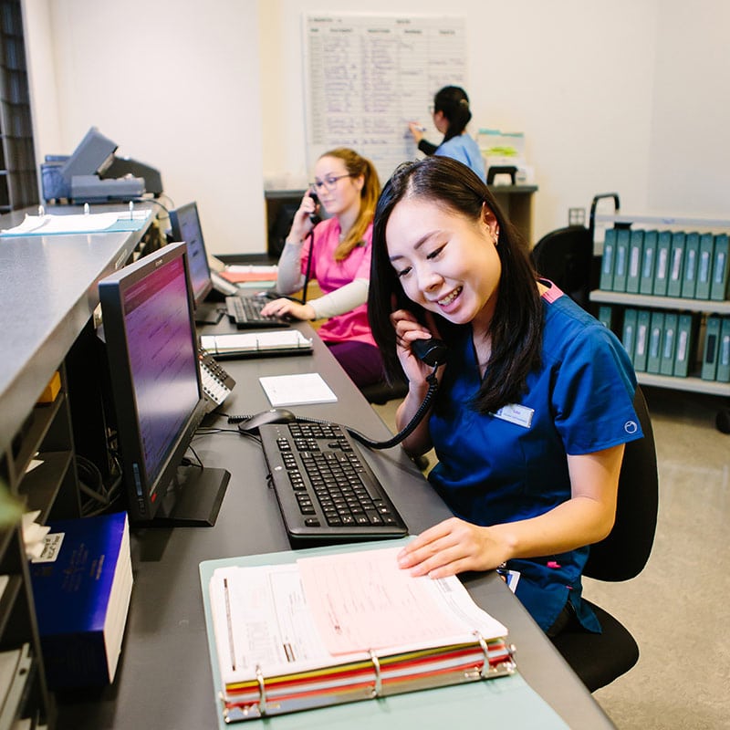 Healthcare unit coordinators or administrative students working at a hospital desk, answering phones and managing patient files and schedules.