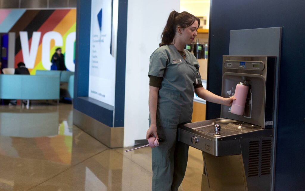 nurse in scrubs refilling a pink water bottle at a water refill station.