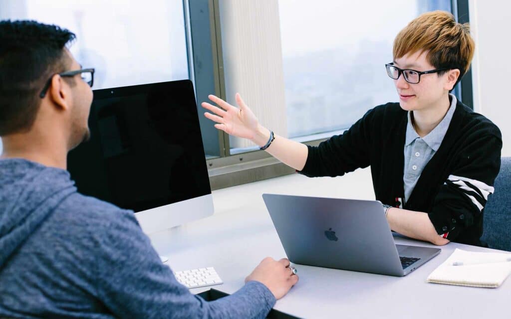 Two people are sitting at a desk indoors, facing each other in discussion.