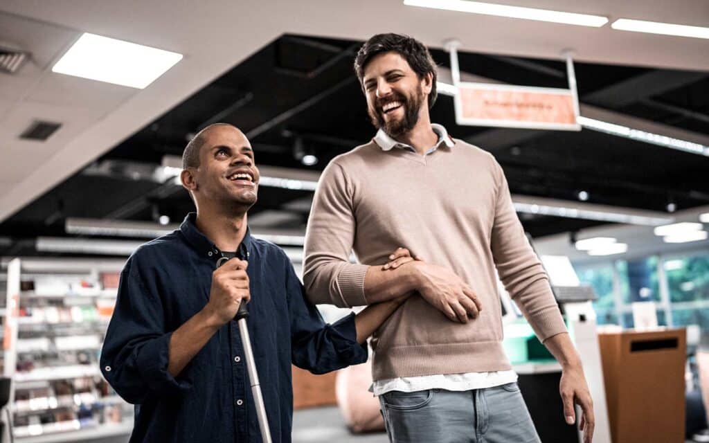 A man with a beard and a sweater guides a smiling blind man holding a cane by walking arm-in-arm through an indoor space.