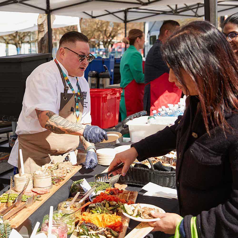 An Indigenous chef with tattoos and wearing a white chef coat serves food from a brightly colored outdoor feast spread to a person.