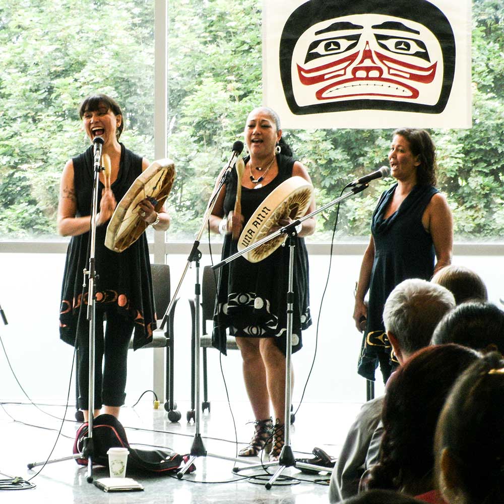 Three Indigenous women stand singing while rhythmically drumming on handheld traditional frame drums in an indoor performance space.