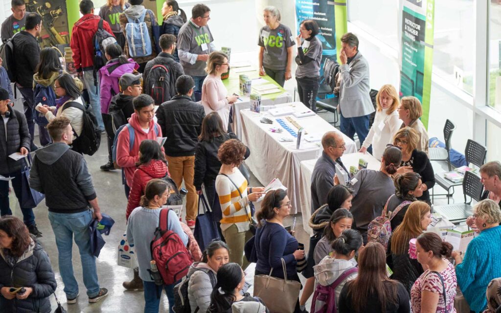 A crowded indoor information session features rows of booths where prospective students interact with staff about various educational programs.