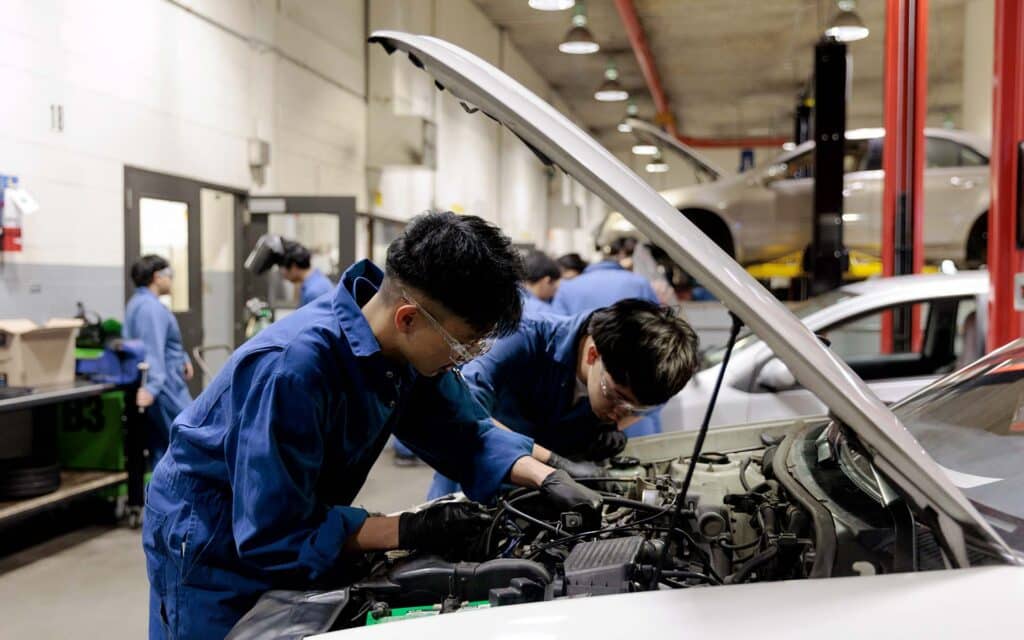 Two mechanics in blue coveralls and safety glasses work together under the open hood of a car in a busy automotive shop.