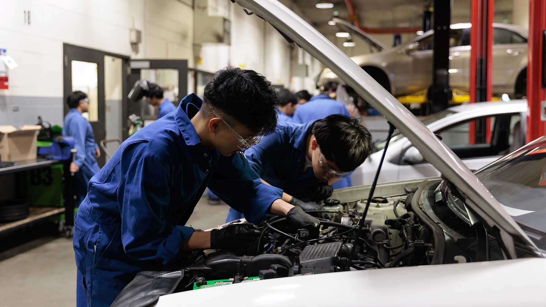 Two mechanics in blue coveralls and safety glasses work together under the open hood of a car in a busy automotive shop.