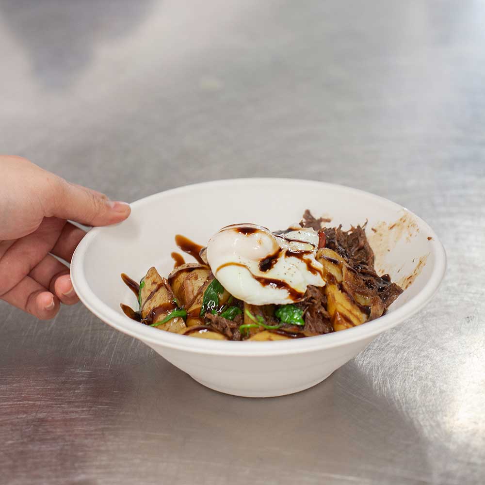 A gourmet bowl of potato wedges topped with shredded beef, fresh greens, and a perfectly poached egg with a balsamic glaze.