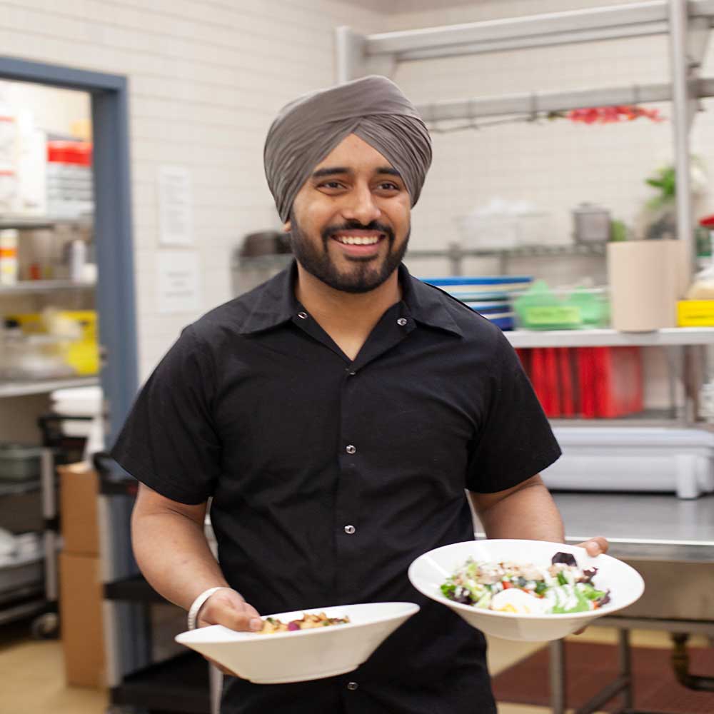 A smiling server in a black uniform carrying two bowls of fresh salad through the kitchen.