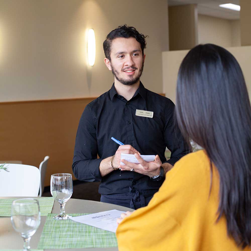 A server in a black shirt taking a guest's order on a notepad at a bistro table.