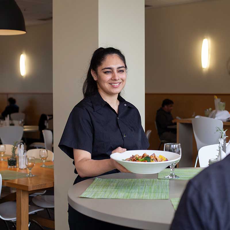 A friendly waitress serving a fresh pasta or salad dish to a customer in a well-lit dining room.