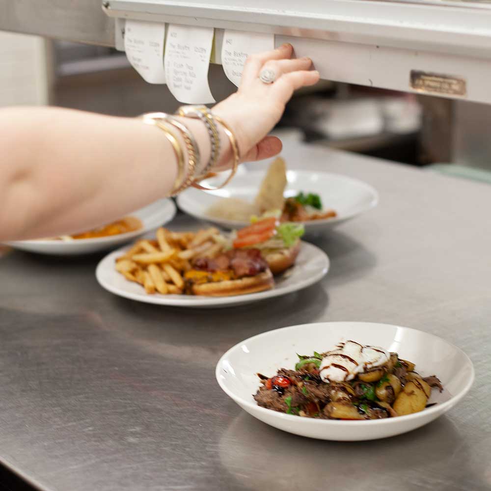 A kitchen staff member reaching for order tickets above a counter lined with prepared gourmet meals.
