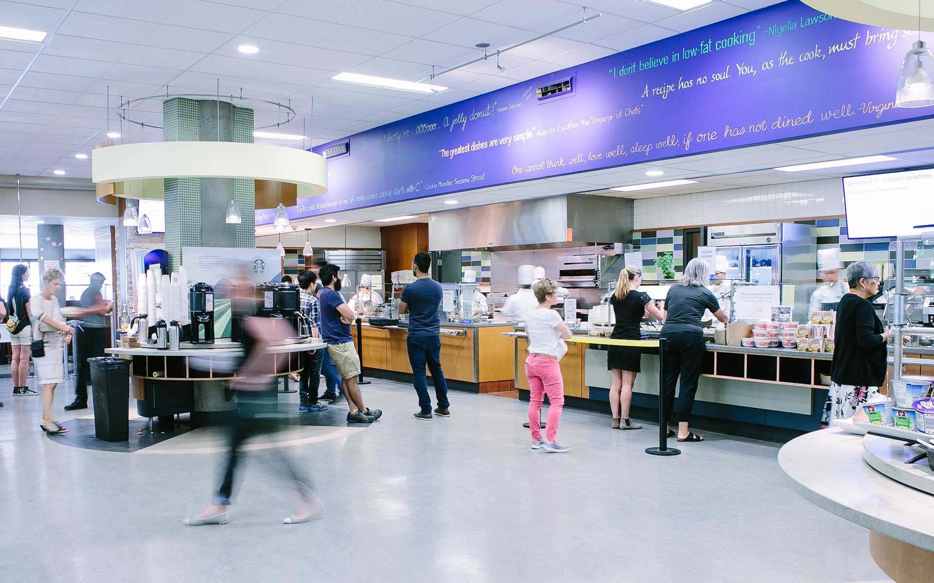Customers wait in line at a modern, bright community cafeteria with purple wall quotes and a student-run kitchen.