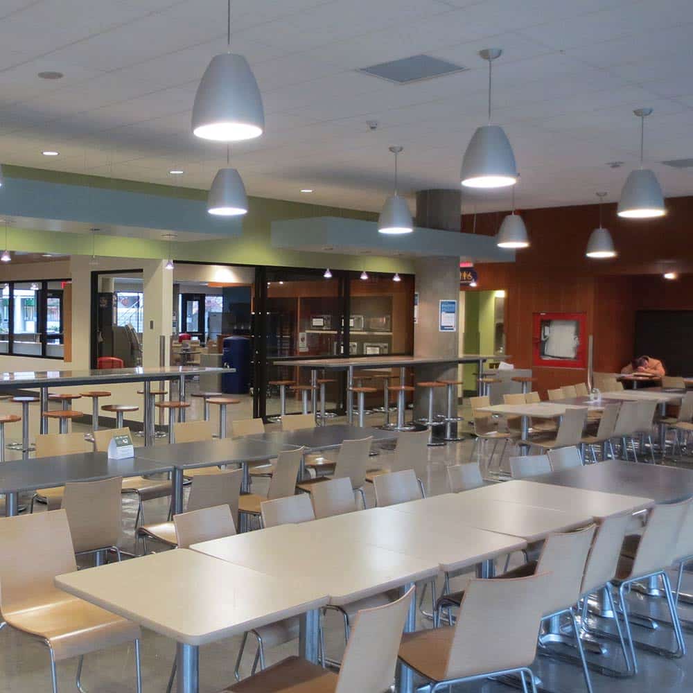 The dining area of a cafeteria featuring rows of clean wooden tables and modern pendant lighting.