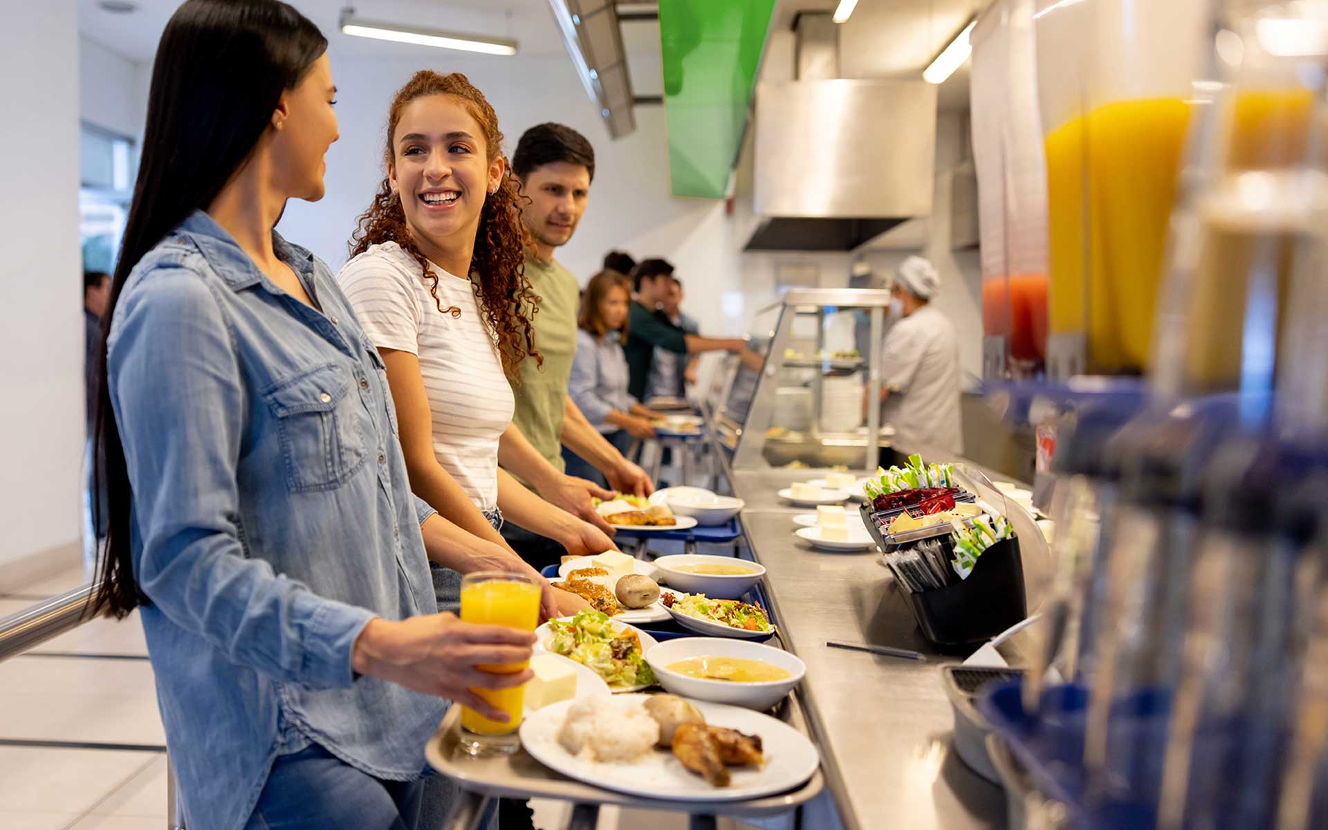 Smiling students holding trays and drinks while moving through a brightly lit cafeteria service line.
