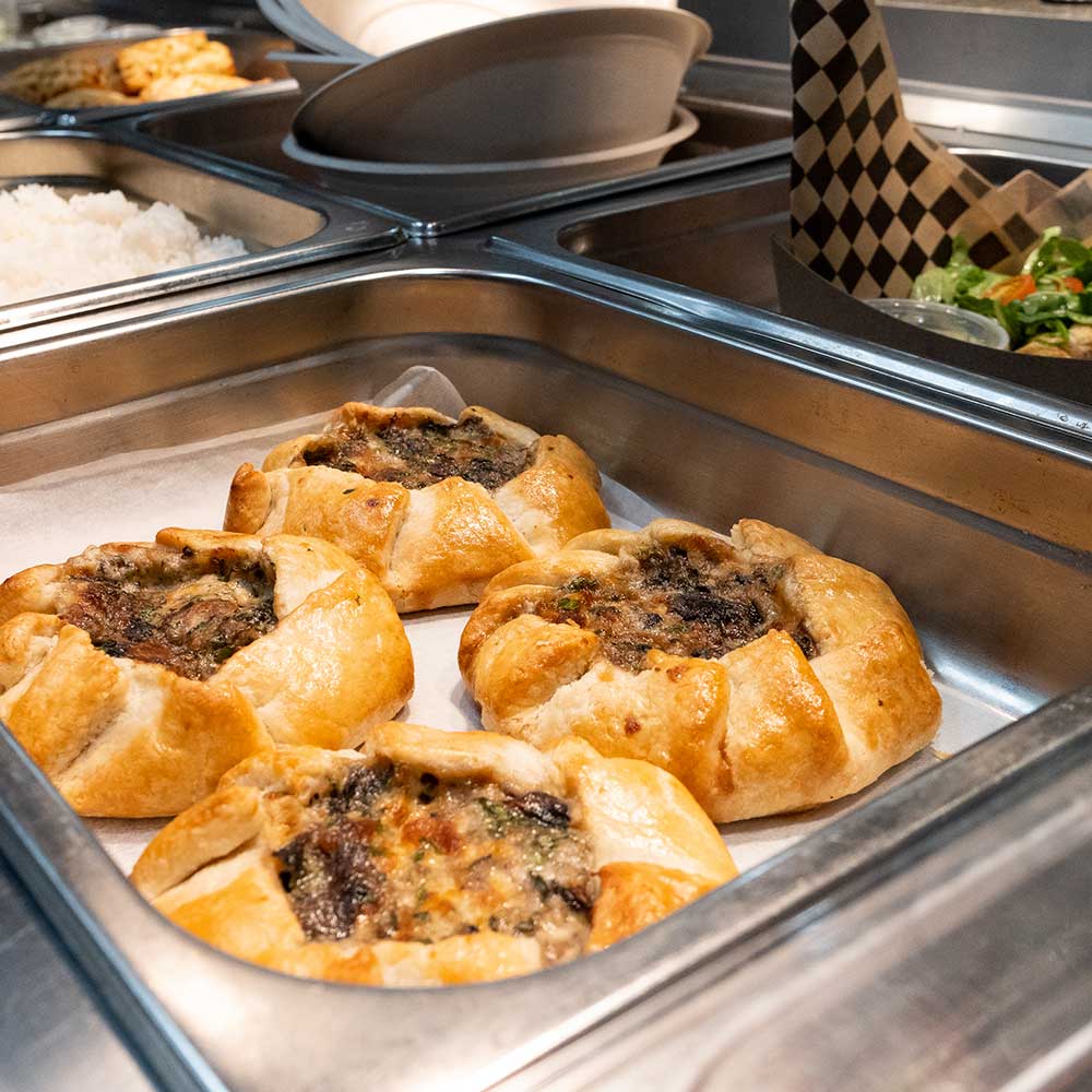 Four golden-brown savory meat pies sitting in a stainless steel warming tray at a food station.