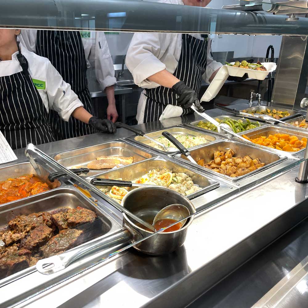 Cafeteria staff in striped aprons serve various hot dishes, including roasted vegetables and meats, from a buffet line."