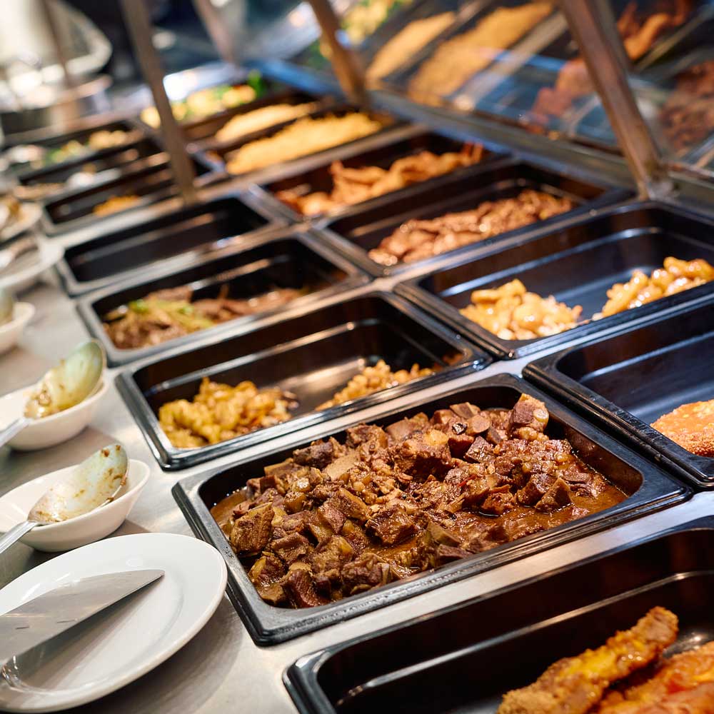 A close-up view of multiple black trays filled with different prepared meat dishes under a heat lamp.