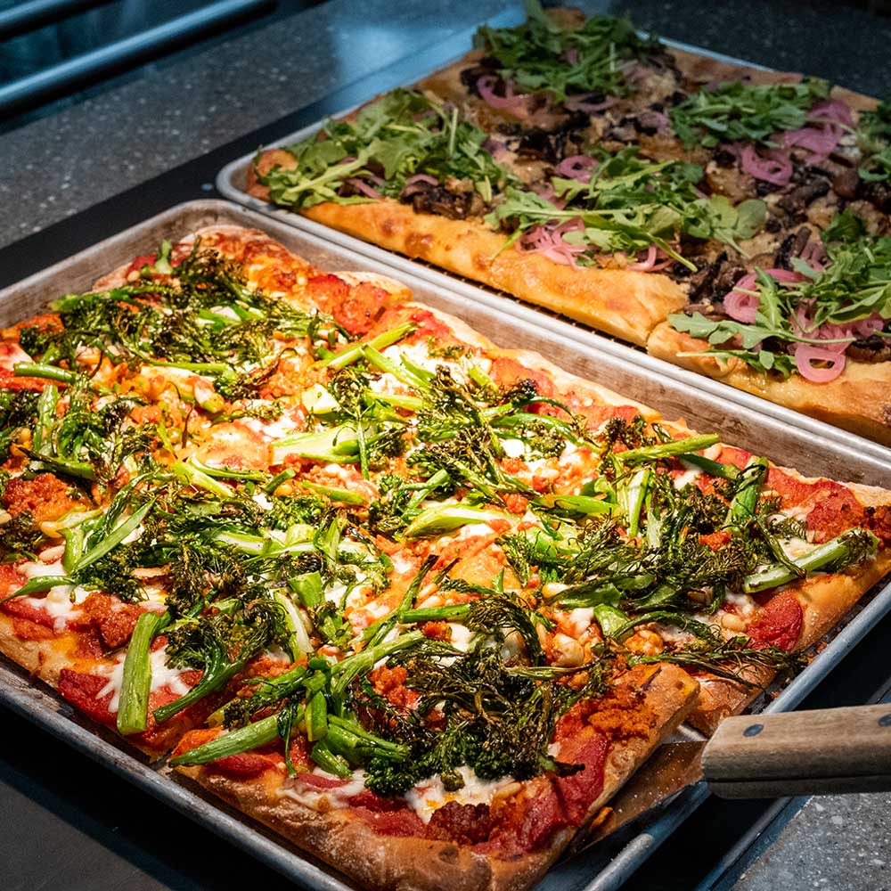 Two large rectangular sheet-pan pizzas topped with fresh greens, broccolini, and red onions.