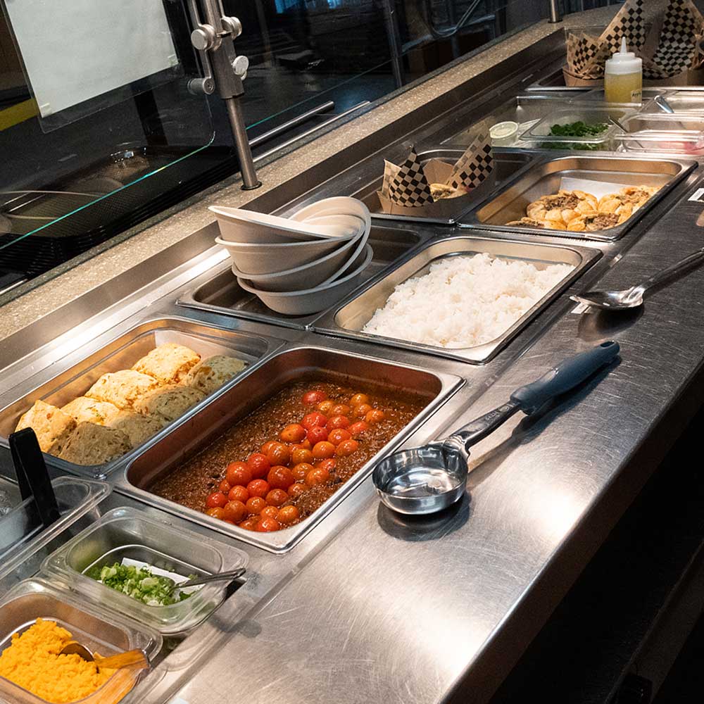 A clean cafeteria service counter with trays of white rice, tomato-based stew, and biscuits.