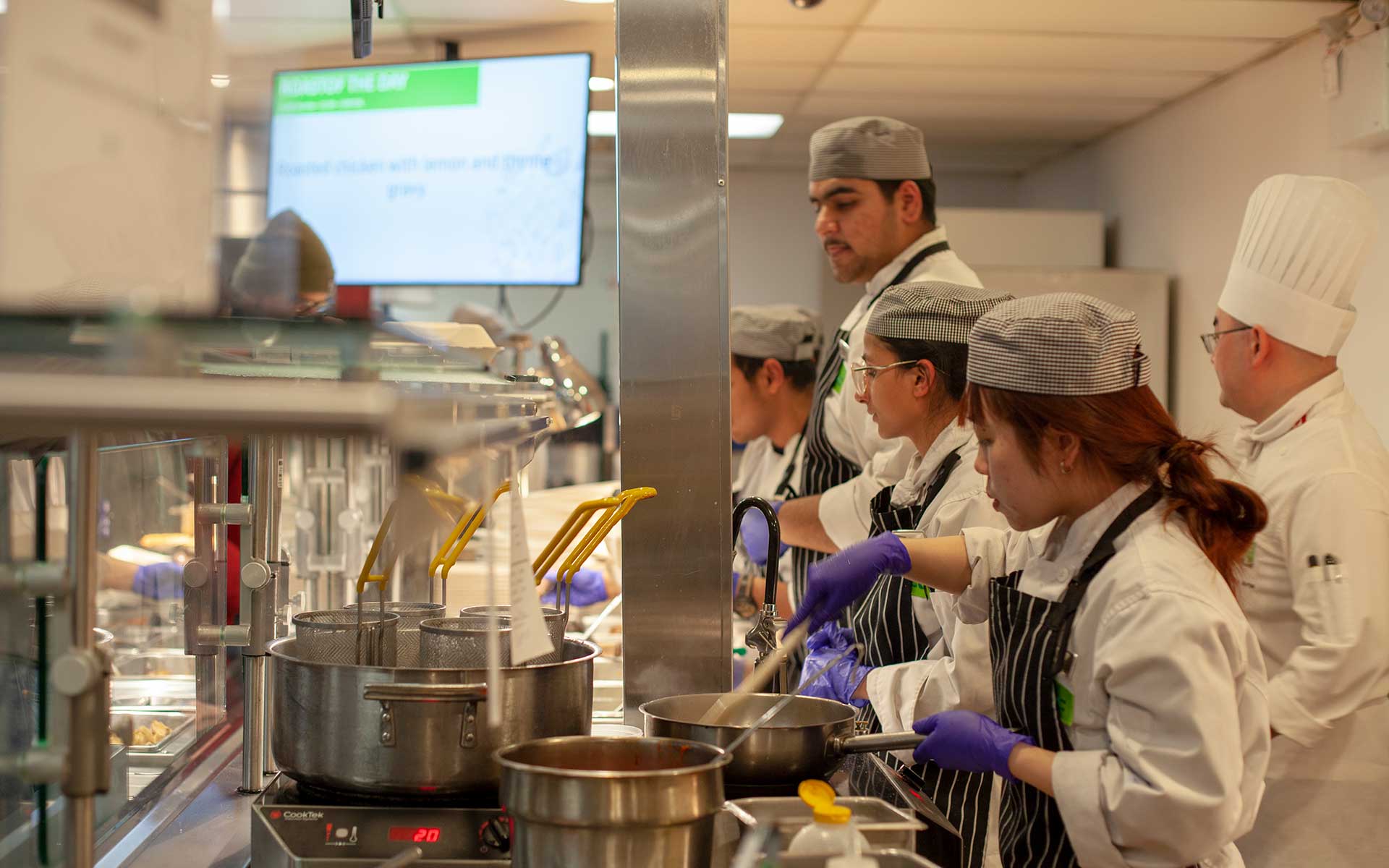 A diverse group of culinary students and chefs wearing uniforms and gloves working at a professional kitchen station.