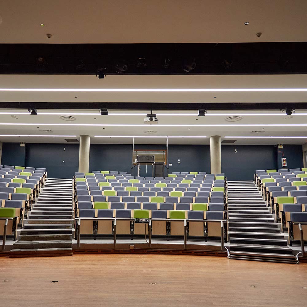 A professional auditorium viewed from the stage, showing tiered seating with a mix of grey and lime-green chairs under bright linear lighting.