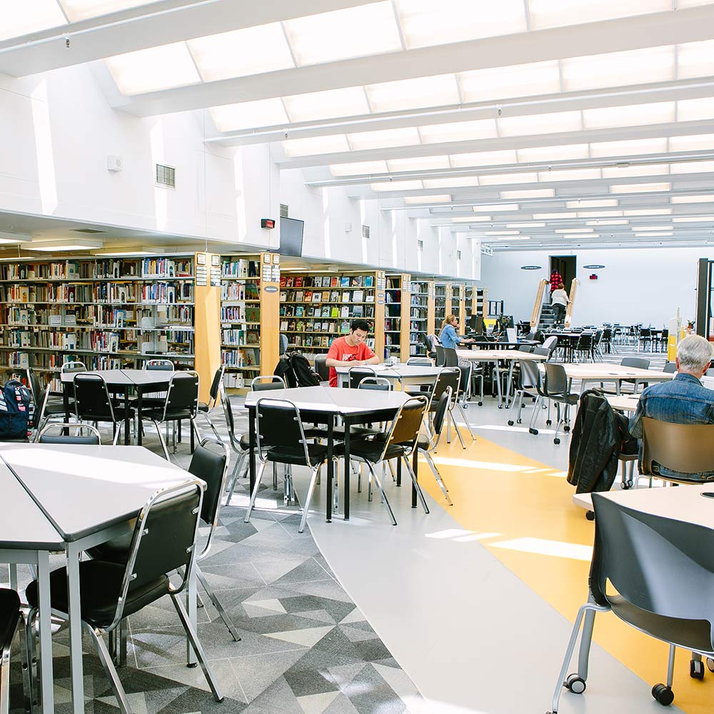 A spacious, well-lit library with yellow floor accents, featuring several round study tables and tall bookshelves filled with various titles.