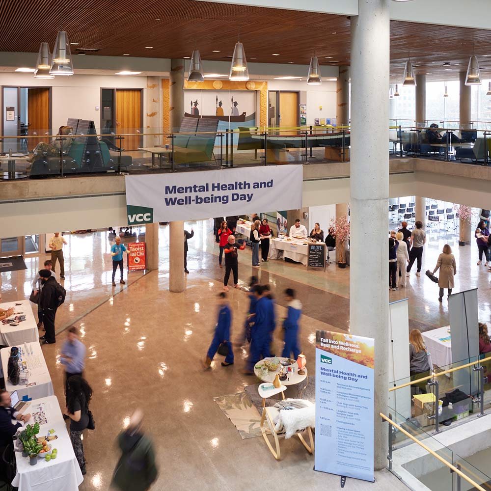 A busy multi-level atrium hosting a "Mental Health and Well-being Day" event with information booths, banners, and people circulating through the space.