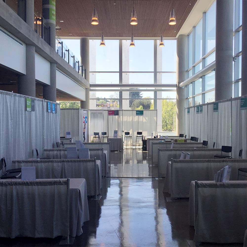 A long, sunlit hall lined with professional draped exhibition booths and tall glass windows overlooking green trees.