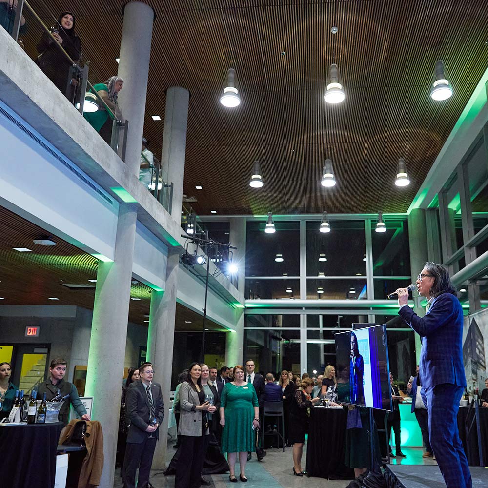 A man in a blue suit speaks into a microphone on a small stage during an evening reception in a modern, glass-walled event space.